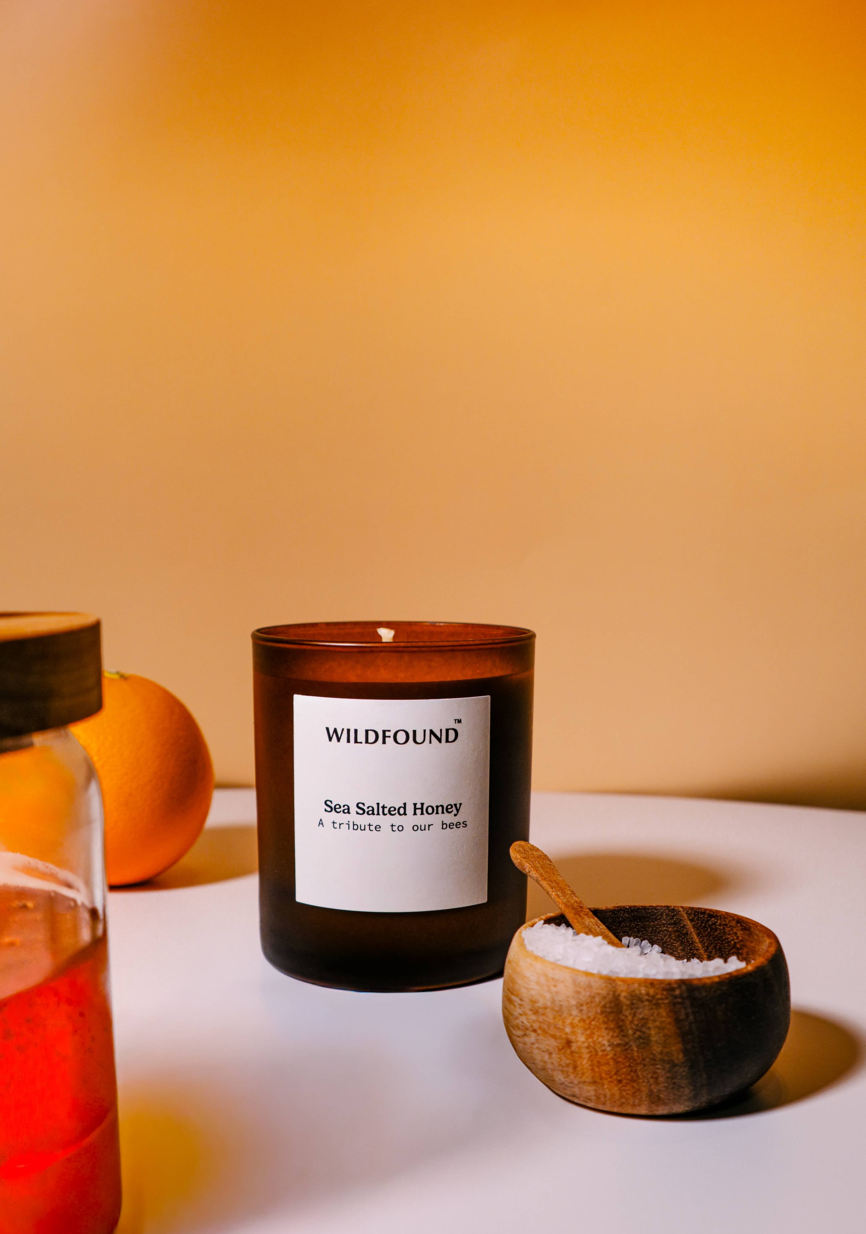 Candle labeled 'Wildfound' with a wooden spoon and container on a white surface with an orange background